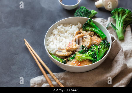 Teriyaki chicken, broccoli and mushrooms stir fry with white rice in bowl on gray concrete background. Asian cuisine. Stock Photo