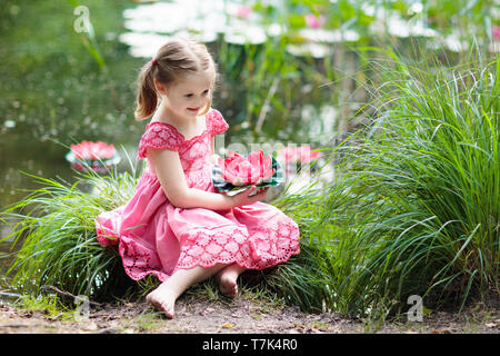 Child sitting at lake shore watching water lily flowers. Little girl holding pink lily flower. Kid looking at lilies on sunny spring day. Kids outdoor Stock Photo