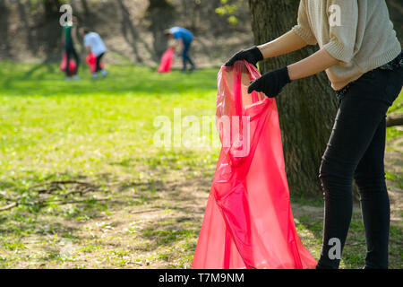 Man and woman volunteer wearing picking up trash and plastic waste in public park. Young people wearing gloves and putting litter into red plastic bag Stock Photo