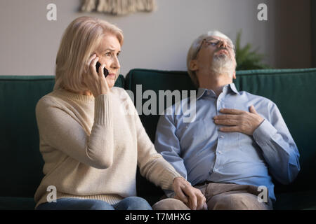 Mature woman calling emergency, grey haired man having heart attack Stock Photo
