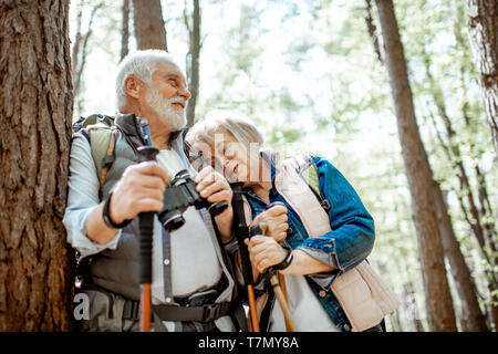 Senior couple resting near the tree while traveling with backpacks and trekking sticks in the forest. Concept of an active lifestyle on retirement Stock Photo