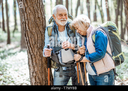 Senior couple resting near the tree while traveling with backpacks and trekking sticks in the forest. Concept of an active lifestyle on retirement Stock Photo