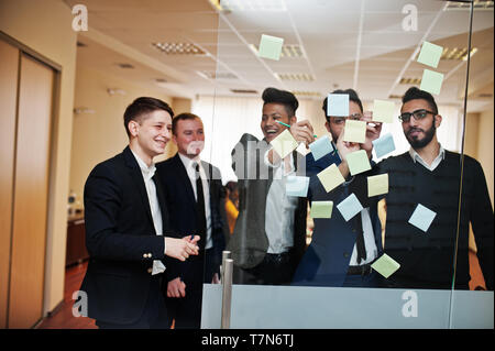Business mans pointing on glass with colorful paper notes. Diverse group of male employees in formal wear using stickers. Stock Photo
