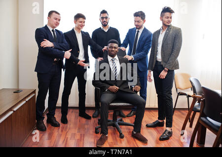 Six multiracial business mans standing at office and roll man on chair. Diverse group of male employees in formal wear having fun. Stock Photo