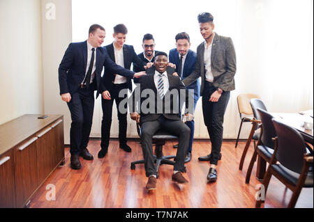 Six multiracial business mans standing at office and roll man on chair. Diverse group of male employees in formal wear having fun. Stock Photo