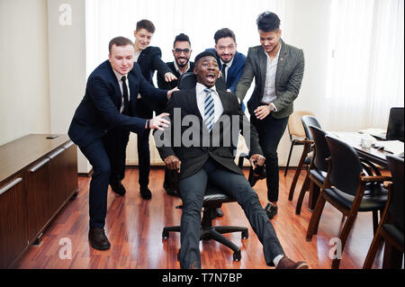 Six multiracial business mans standing at office and roll man on chair. Diverse group of male employees in formal wear having fun. Stock Photo