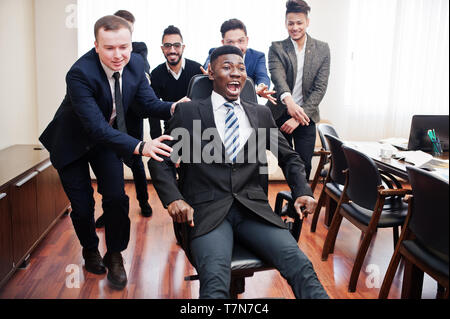 Six multiracial business mans standing at office and roll man on chair. Diverse group of male employees in formal wear having fun. Stock Photo