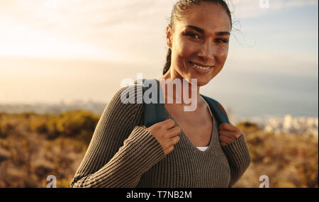 Happy woman carrying a bag walking through field. Young female on country hike, looking at camera and smiling. Stock Photo