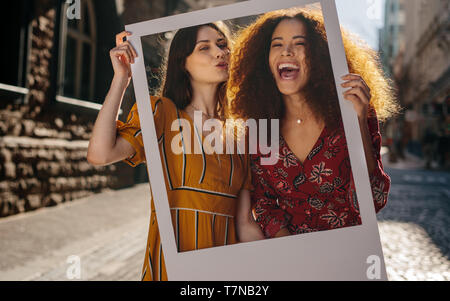 Excited young women holding a empty picture frame while standing outdoors. Multi-ethnic female traveler friends with photo frame. Stock Photo