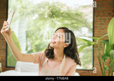 Beautiful young woman is holding a mop, using a smartphone and smiling while cleaning her house Stock Photo