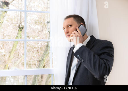 Closeup of serious young man businessman in business suit and tie by window in room listening talking on phone thinking Stock Photo