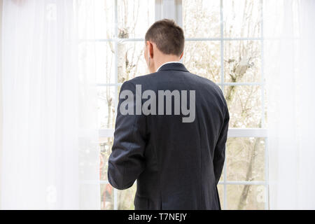 Back of young man businessman in suit standing by window in home listening to earbuds headphones listening to music Stock Photo