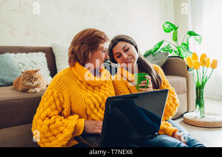 Senior mother and her adult daughter using laptop at home while drinking tea. Mother's day concept. Stock Photo