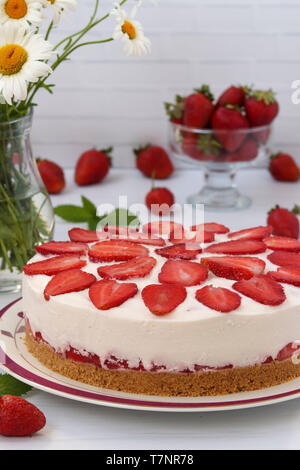 Cake with strawberries without baking, with a base of biscuits, localed vertically on a plate on a white background, a bouquet of daisies in the backg Stock Photo