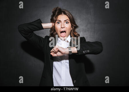 Portrait of a shocked young businesswoman wearing formal suit standing isolated over black background, looking at wrist watch Stock Photo