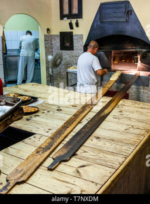 Two long, well-worn wooden peels lie in the foreground on a work table in a traditional bakery in Dobrich, Bulgaria. In the background, a baker slides gevrek rings into the oven while a second baker works in the adjoining kitchen. Round, already baked pastries lie on a tray to the left. The workshop probably belongs to the Stariyat Dobrich open-air museum (Old Dobrich), which brings together more than 30 craft workshops. Stock Photo