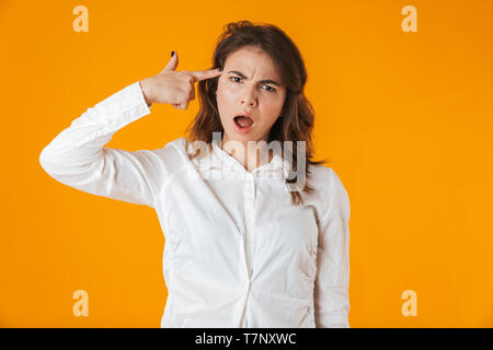 Portrait of a mad young woman wearing white shirt standing isolated over yellow background, pointing finger at her head Stock Photo