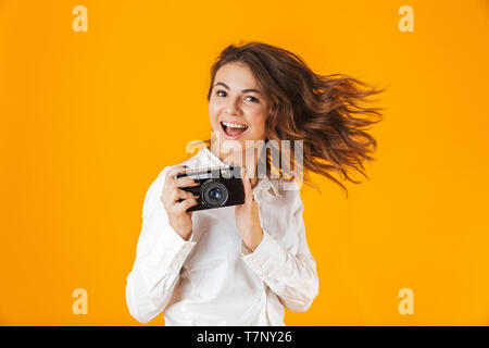 Cheerful young woman casualy dressed standing isolated over yellow background, taking photo with camera Stock Photo