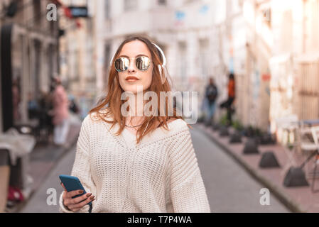 Beautiful attractive young trendy girl in jumper and jeans with headphones smartphone and sunglasses listen music while walking in crowd
