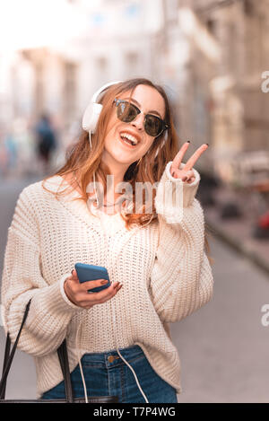 Beautiful attractive young trendy girl in jumper and jeans with headphones smartphone and sunglasses makes peace sign with her hands while walking at  Stock Photo