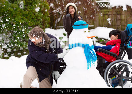 Caucasian father having fun  playing snowball fight with biracial children outdoors in winter. Young boy is disabled in wheelchair. Stock Photo