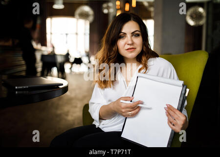 Young woman sits in cafe at a table, holding pen, reading documents. A businesswoman signs a contract. Startup, entrepreneur working. Stock Photo