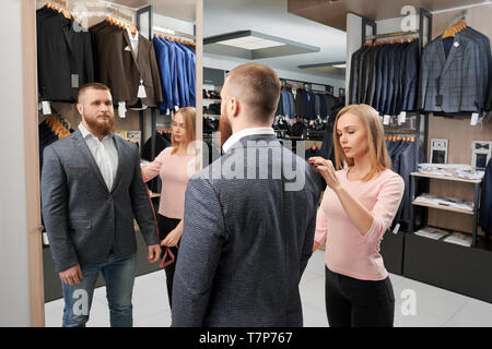 View from back of serious male client trying on new jacket and looking mirror while pretty female assistant measuring length of sleeve in clothes shop. Concept of style and fitting. Stock Photo