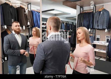 View from side of bearded man trying new white shirt and grey casual jacket in shop. Smiling man standing in front of mirror while female tailor measuring length of sleeve. Concept of fitting. Stock Photo