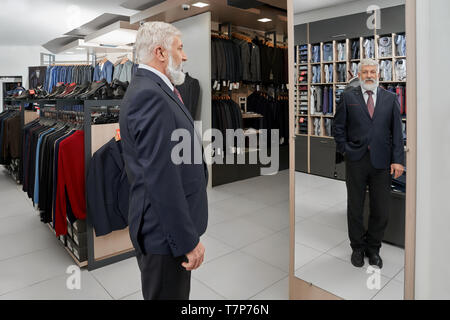 Smiling elder man wearing official smart suit looking at mirror in big clothes boutique. Bearded businessman choosing and purchasing outfit for holiday. Concept of fitting and elegance. Stock Photo