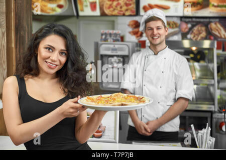 Happy female customer keeping tasty pizza in hands and posing in cafe. Attractive brunette looking at camera and smiling while professional male cook standing at background. Concept of pizzeria. Stock Photo