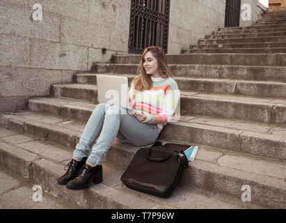 Attractive trendy teen student girl or young college woman working on laptop on the Internet, blogging, chatting or on steps in urban background In On Stock Photo