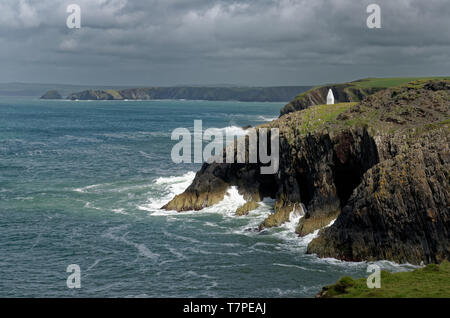 Dramatic cliffs and a white stone navigation marker in the sunlight on the Pembrokeshire Coast Path at the entrance to Porthgain harbour, Wales Stock Photo