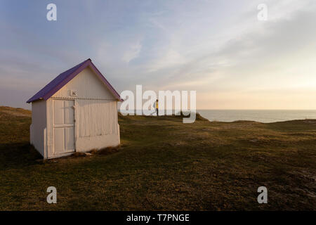 Woman with a backpack in yellow jacket walking on grass at row of white wooden beach huts with colorful roofs, Gouville-sur-Mer, Normandy, France
