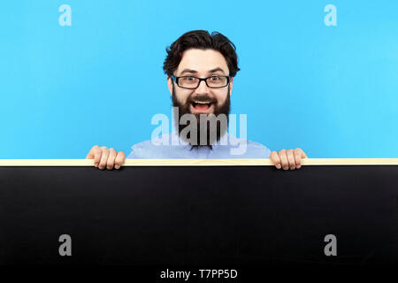cheerful bearded man in glasses,  showing a  chalk board over blue background, concept of  positive teacher Stock Photo