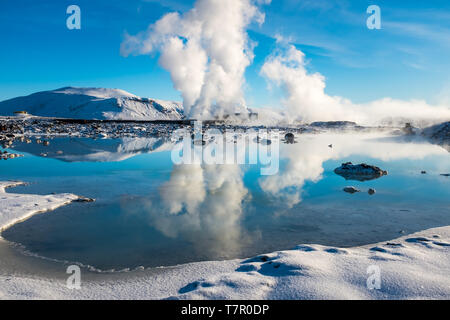 Steam from a geothermal plant shoots skywards against a bright blue sky and reflected in a lake beside the Blue Lagoon Stock Photo