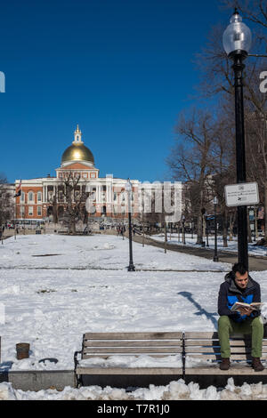 Boston, MA, USA, February, 8, 2016: A young man perches on the back of outdoor bench reading a book on a chilly snow covered park with a back drop of the Massachusetts Capitol Building against a bright blue sky Stock Photo
