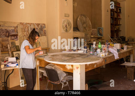 Spello, Umbria, Italy, July, 31, 2017: A young female in her studio working on a large round art piece, paints and brushes are scattered all around her. Stock Photo
