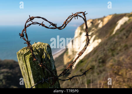 Barbed wire on a fence post shaped into a heart with a backdrop​ of white cliffs in Devon, against a clear blue sky Stock Photo