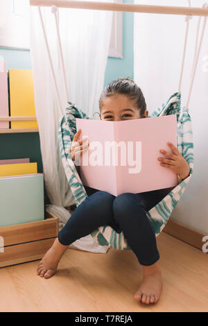 vertical photo of a sweet happy little girl reading a book in her room near the window, funny lovely child having fun in kids room Stock Photo