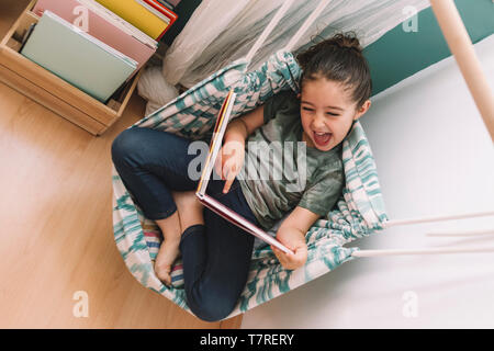 little girl laughs out loud reading a book at home near the window, funny lovely child having fun in her kids room, copy space for text Stock Photo