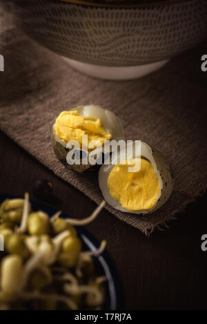 Vertical photo of quail egg broken to two halves. Nice yolk with bright yellow color is inside. Bowl with next eggs is next to broken egg. Eggs are on Stock Photo