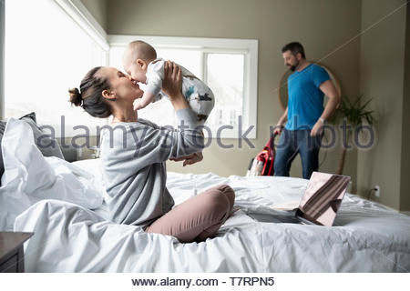 Affectionate mother playing with baby son on bed while husband vacuums in background