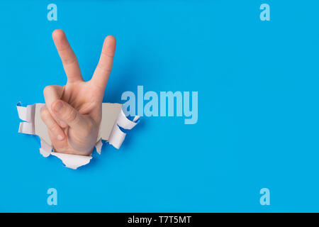 Male hand tearing through blue paper making peace sign with fingers and copt space for text. Stock Photo