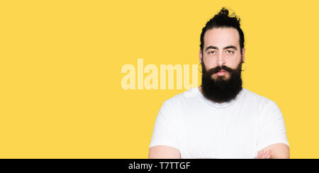 Young hipster man with long hair and beard wearing casual white t-shirt skeptic and nervous, disapproving expression on face with crossed arms. Negati Stock Photo