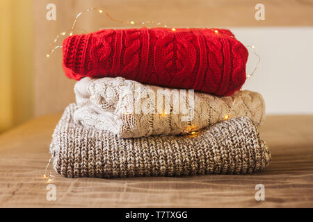 A pile of knitted sweaters on the wood bed decorated with lights. Warm comfortable clothes. Stock Photo