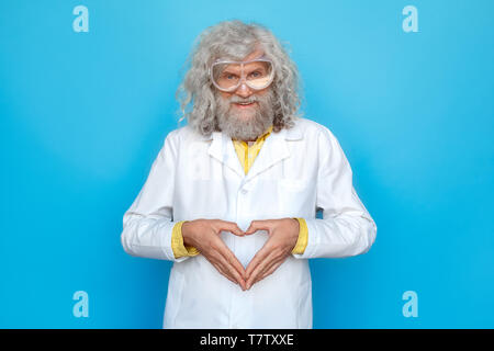Old long-haired man wearing doctor's gown and goggles studio isolated on blue wall standing looking camera smiling friendly making heart shape with ha Stock Photo