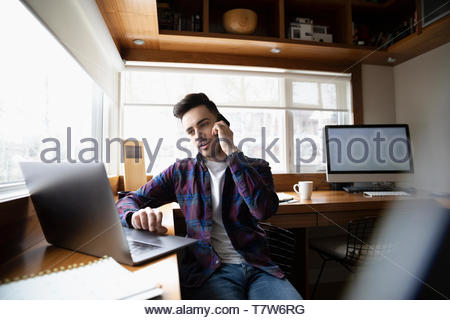 Young man working at home, talking on cell phone at laptop in home office