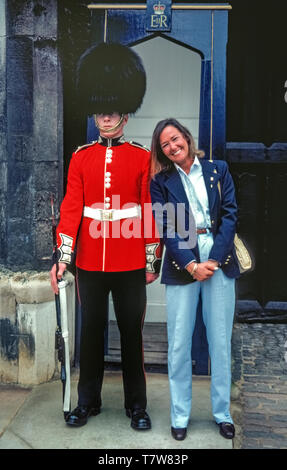 A Queen's Guard in the traditional red tunic and black bearskin hat keeps his composure as a female tourist snuggles up to him for a souvenir photograph in front of his sentry box at St. James's Palace in London, England. Since 2014 the guards and their sentry boxes have been relocated inside gates to the palace and tourists are no longer able to pose beside them. Also, the guards nowadays carry the British Army's standard-issue automatic assault rifles instead of the type of gun seen in this historical 1981 photograph. Stock Photo