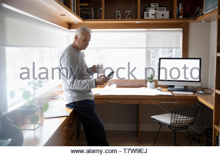 Senior man working from home, drinking coffee and using smart phone in home office