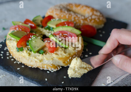 Bagel with hummus, avocado slices, spices, green onion, tomatoes cherry and sesame seeds on the black stone desk. Hand with knife with hummus. Close u Stock Photo